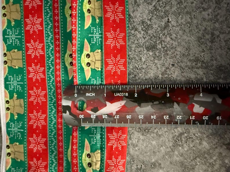 Puede incluir: Una secci&oacute;n de tela con un patr&oacute;n festivo con tem&aacute;tica navide&ntilde;a. El dise&ntilde;o presenta rayas rojas y verdes alternas con motivos de copos de nieve y Grogu. Una regla muestra que la tela mide unos 5 cm de ancho.