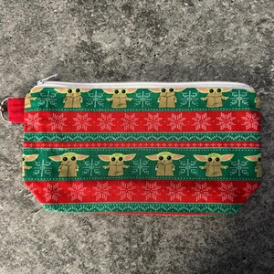 Puede incluir: Una bolsa rectangular con cremallera y un estampado festivo. El dise&ntilde;o presenta un patr&oacute;n repetitivo de un personaje de una popular franquicia de ciencia ficci&oacute;n, copos de nieve y rayas en rojo y verde. Se adjunta un lazo de tela roja y un cierre plateado.