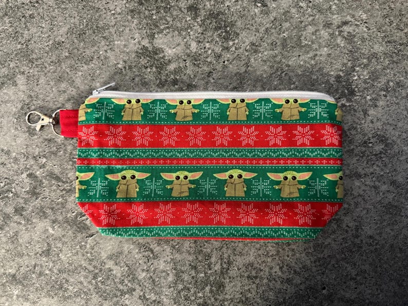 Puede incluir: Una bolsa rectangular con cremallera y un estampado festivo. El dise&ntilde;o presenta un patr&oacute;n repetitivo de un personaje de una popular franquicia de ciencia ficci&oacute;n, copos de nieve y rayas en rojo y verde. Se adjunta un lazo de tela roja y un cierre plateado.