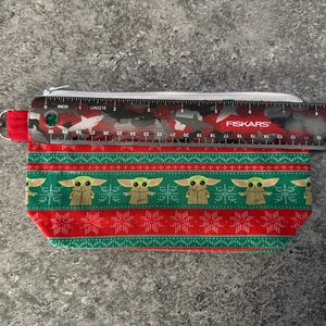Puede incluir: Una bolsa rectangular con cremallera con un patr&oacute;n rojo y verde que presenta una imagen repetida de Grogu de The Mandalorian. La bolsa mide aproximadamente 18 cm de largo. Una regla est&aacute; colocada encima.