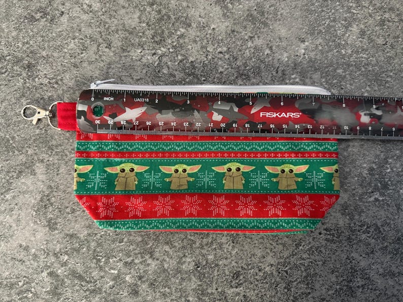 Puede incluir: Una bolsa rectangular con cremallera con un patr&oacute;n rojo y verde que presenta una imagen repetida de Grogu de The Mandalorian. La bolsa mide aproximadamente 18 cm de largo. Una regla est&aacute; colocada encima.