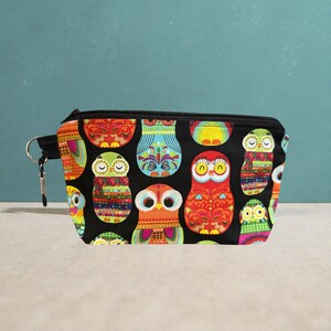 Peut inclure: Une pochette noire zippée ornée d'un motif de hiboux colorés. Les hiboux sont de différentes couleurs, notamment rouge, vert et orange, avec des motifs complexes. La pochette a une fermeture éclair noire et un petit anneau argenté sur le côté.