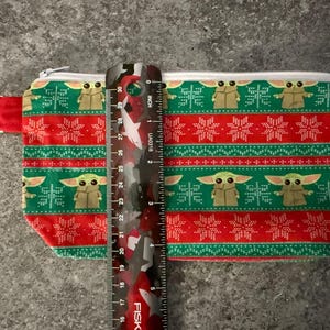 Puede incluir: Una bolsa rectangular con cremallera con un estampado festivo con tem&aacute;tica navide&ntilde;a. El dise&ntilde;o presenta un patr&oacute;n repetitivo de un personaje verde, copos de nieve y rayas rojas. La bolsa mide aproximadamente 25 cm de largo.