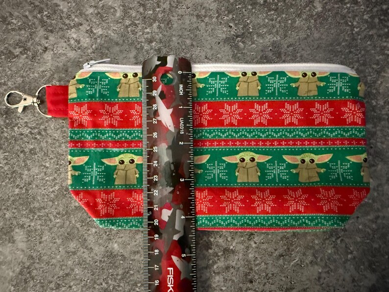 Puede incluir: Una bolsa rectangular con cremallera con un estampado festivo con tem&aacute;tica navide&ntilde;a. El dise&ntilde;o presenta un patr&oacute;n repetitivo de un personaje verde, copos de nieve y rayas rojas. La bolsa mide aproximadamente 25 cm de largo.