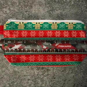 Puede incluir: Estuche rectangular con cierre de cremallera, con un dise&ntilde;o festivo. El estuche mide aproximadamente 20 cm de largo y tiene un patr&oacute;n rojo y verde con motivos de copos de nieve y personajes. Se adjunta un cierre plateado.