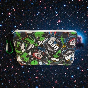 May include: A small, zippered pouch featuring a collage of green alien and UFO-themed graphics on a black background. The design includes the words "UFO Area" and "Space Explore". A green carabiner clip is attached to the side.
