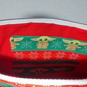 Puede incluir: Vista interior de una bolsa de tela roja con cremallera blanca. La bolsa presenta tres secciones de tela con un estampado verde y rojo. La tela verde tiene una imagen repetida de un personaje de dibujos animados.