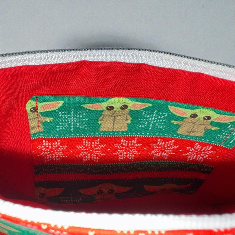 Puede incluir: Vista interior de una bolsa de tela roja con cremallera blanca. La bolsa presenta tres secciones de tela con un estampado verde y rojo. La tela verde tiene una imagen repetida de un personaje de dibujos animados.