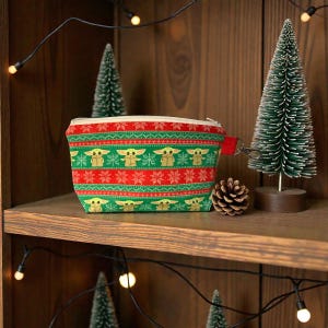 Puede incluir: Peque&ntilde;o neceser rectangular con cierre de cremallera. La bolsa presenta un patr&oacute;n festivo de rayas verdes y rojas con una imagen repetida de un personaje. Una correa de mu&ntilde;eca roja est&aacute; unida a la cremallera. La bolsa se exhibe en un estante de madera.