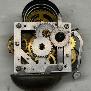 May include: Close-up of a vintage clock mechanism. The image shows a complex arrangement of gears in gold and silver tones, along with a dark bell and a coiled spring. The intricate details highlight the craftsmanship of the clock's internal workings.