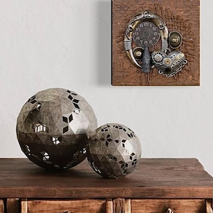 May include: Two metallic spheres with a geometric faceted surface and cut-out patterns, paired with a square wall art piece featuring a circular clock face, a heart-shaped element, and various mechanical components on a textured brown background.