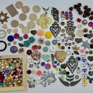 May include: A collection of colorful and metallic embellishments, including beads, buttons, charms, and wooden shapes. The items are arranged on a white surface and include a variety of colors, textures, and designs.