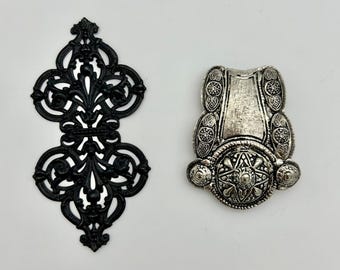 May include: Two decorative metal embellishments. One is black and features a filigree design. The other is silver and has a more ornate design with a central star.