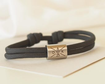 May include: A dark gray paracord bracelet with a silver charm engraved with the letters "A D K" and a heart symbol, along with the number "25".