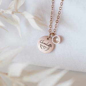 Op de afbeelding: Een ros&eacute;gouden ketting met een ronde hanger waarop "Charlotte" staat met engelenvleugels en een kleine, heldere kristallen hanger.