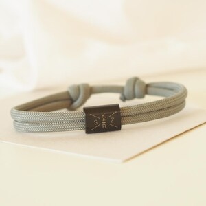 May include: A grey paracord bracelet with a black metal bead engraved with the letters "KSZB" and an anchor symbol.