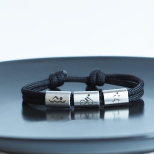 May include: A black paracord bracelet with three silver beads. Each bead features a different symbol: a swimmer, a cyclist, and a runner.