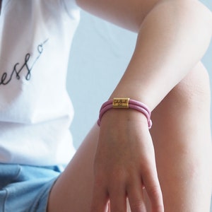 Könnte beinhalten: Ein pinkfarbenes Armband aus Kordel mit einem goldenen Anhänger, auf dem "Schulland 2023" steht.