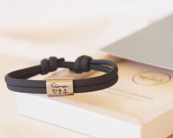 May include: A black paracord bracelet with a silver charm engraved with the name "Simon" and symbols of a heart, cross, and anchor.
