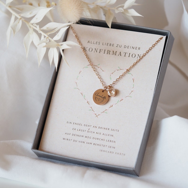 Op de afbeelding: Een gouden ketting met een klein rond hangertje waarop "Charlotte" staat met een vlinderontwerp. De ketting zit in een doosje met een kaartje waarop "Alles Liebe zu deiner Konfirmation" staat en een citaat over een engel die over je waakt.