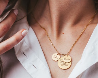 Op de afbeelding: Gouden ketting met drie ronde hangers. De grootste hanger zegt "Sisters" met een hartje. De twee kleinere hangers zeggen "Julia" en "Carina".