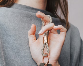 May include: A person is holding a grey rope keychain with a gold metal ring and a gold metal tag that says "Pandini". A silver key is attached to the keychain.