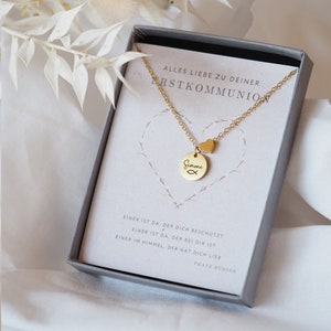 Könnte beinhalten: Eine goldene Halskette mit einem kleinen Herz-Anhänger und einem runden Anhänger mit dem Namen "Simone" und einem Fischsymbol. Die Halskette befindet sich in einer weißen Schachtel mit der Aufschrift "Alles Liebe zu deiner Erstkommunion" und einem Zitat von Franz Bübner.