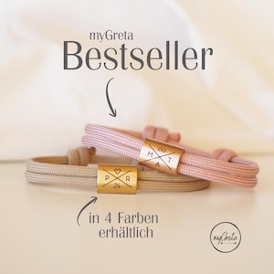 Könnte beinhalten: Zwei Kordelarmbänder mit goldfarbenen Metallverschlüssen. Eines ist hellrosa, das andere beige. Die Verschlüsse haben eingravierte Initialen und Symbole. Der Text "Bestseller" und "in 4 Farben erhältlich" sind ebenfalls sichtbar.