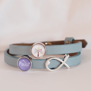Könnte beinhalten: Ein hellblaues Lederarmband mit drei Anhängern: einem silbernen Unendlichkeitszeichen, einem lila Anhänger mit dem Namen "Paula" in weißen Buchstaben und einem runden Anhänger mit einem bunten Baummotiv.