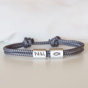 May include: Grey rope bracelet with two silver beads. One bead is engraved with the name "PAUL" and the other with a fish symbol.
