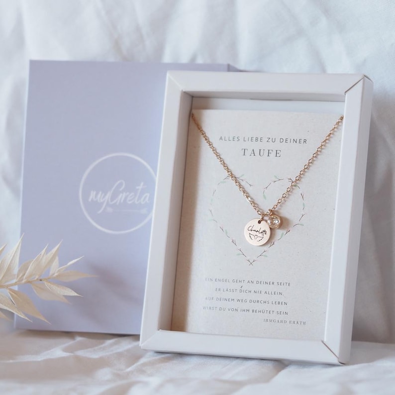 Op de afbeelding: Een witte cadeaudoos met een gouden ketting erin. De ketting heeft een klein rond hangertje met het woord "Charl" erop gegraveerd. De doos heeft een witte kaart met de tekst "Alles Liebe zu deiner Taufe" en een gedicht over engelen.