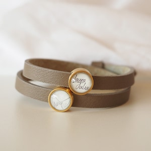Op de afbeelding: Een bruin leren armband met dubbele wikkel en twee goudkleurige bedels. Een bedel heeft een handgeschreven inscriptie "Laura Julia" en de andere bedel heeft een lijntekening van twee handen die elkaar vasthouden.