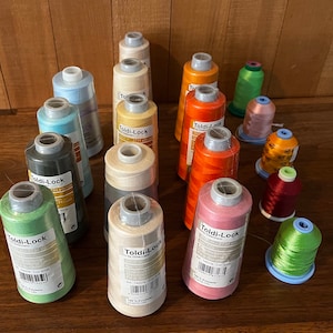 May include: A collection of 15 spools of thread in various colors, including green, pink, red, orange, beige, and blue. The spools are labeled "Toldi-Lock" and have different numbers and descriptions.