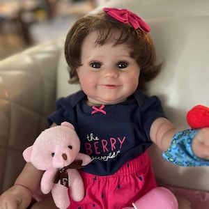 May include: A baby doll with brown hair and brown eyes is wearing a blue shirt with the text "the berry sweetest" on it, pink shorts with a watermelon print, and a pink bow in her hair. She is holding a pink teddy bear and a bottle.