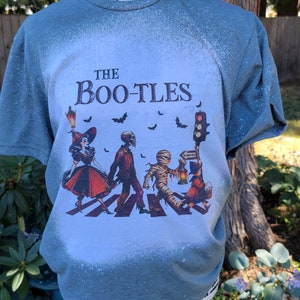 May include: A blue t-shirt with a bleach-splattered design featuring a Halloween-themed graphic of a witch, skeleton, mummy, and a ghost crossing a street with the text "THE BOO-TLES".