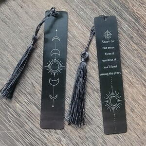 May include: Two black metal bookmarks with black tassels. One bookmark features a sun and moon design with the quote "Shoot for the moon. Even if you miss it, you'll land among the stars."