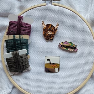 May include: A wooden embroidery hoop with three spools of embroidery floss in shades of purple, teal, and gray. Three enamel pins are displayed inside the hoop: a brown Highland cow, a glass with a Nessie monster inside, and a pink and green pin with the text "Evergreen".