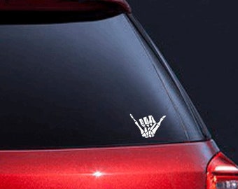 May include: White vinyl decal of a skeleton hand making the shaka sign, a popular Hawaiian gesture of peace, aloha, and good vibes. The decal is on the back window of a red car.