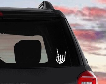 May include: A white skeleton hand sticker with the index and pinky fingers extended, forming a rock and roll hand gesture. The sticker is on the back window of a black car.