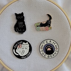 May include: Four enamel pins featuring black cats. One pin features a black cat with the text "Good Luck". Another pin features a black cat curled up on a bed of pink and green flowers. The third pin features a white cat with the text "Fat Cat. Fan Club." The fourth pin features a black paw print with the text "Team Toe Beans".