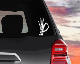 May include: A white skeleton hand making the 'okay' sign. The hand is on a black car with a red taillight.