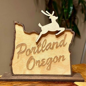 May include: Wooden sign shaped like the state of Oregon with a deer silhouette jumping over the top. The sign says "Portland Oregon" in a cursive font.