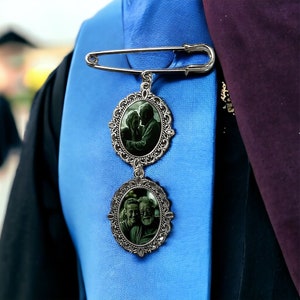 May include: A silver brooch with two oval-shaped photo charms. The top charm features a couple embracing, while the bottom charm shows two people smiling. The brooch is attached to a safety pin.