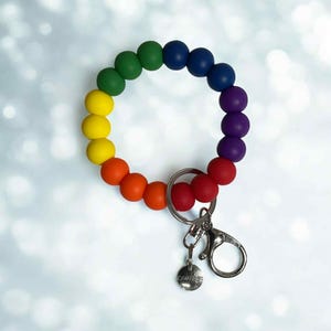 May include: A rainbow-colored silicone bead keychain wristlet. The beads are arranged in a circular pattern, transitioning from red to orange, yellow, green, blue, and purple. It includes a silver-tone clasp and a small charm with the word "Courage".