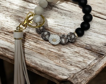 May include: A white and black beaded keychain with a white daisy charm and a white tassel. The beads are made of different materials, including marble, leopard print, and solid black.