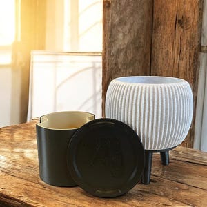 May include: A white planter with a ribbed texture and three black legs. The planter is sitting on a wooden surface. A black lidded container with a cream interior is next to the planter.