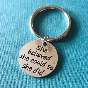 May include: A silver-colored keychain with a round pendant. The pendant has the words "She believed she could so she did" engraved in a black, bold font. The keychain is on a light blue background.