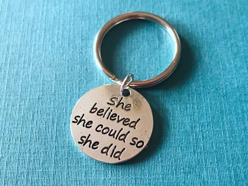 May include: A silver-colored keychain with a round pendant. The pendant has the words "She believed she could so she did" engraved in a black, bold font. The keychain is on a light blue background.