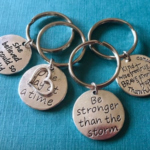 May include: A collection of silver keychains with inspirational quotes. The keychains have round pendants with phrases like "She believed she could so she did" and "Be stronger than the storm". The background is a solid teal color.