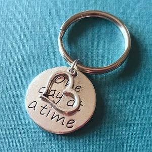 May include: A silver-colored keychain with a round charm and a heart-shaped cutout. The charm is inscribed with the words "One day at a time" in a handwritten style font. The keychain is on a blue background.